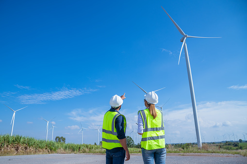 Engineer wearing uniform ,helmet hold document inspection work in wind turbine farms rotation to generate electricity energy. Green ecological power energy generation wind sustainable energy concept.