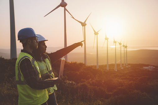 Young electrical engineer woman and business man standing in front of wind turbines checking and working about technical problems and writes the results of measurements with laptop pc in wind power plant electric energy station. xxxl size taken with canon 5d mIV